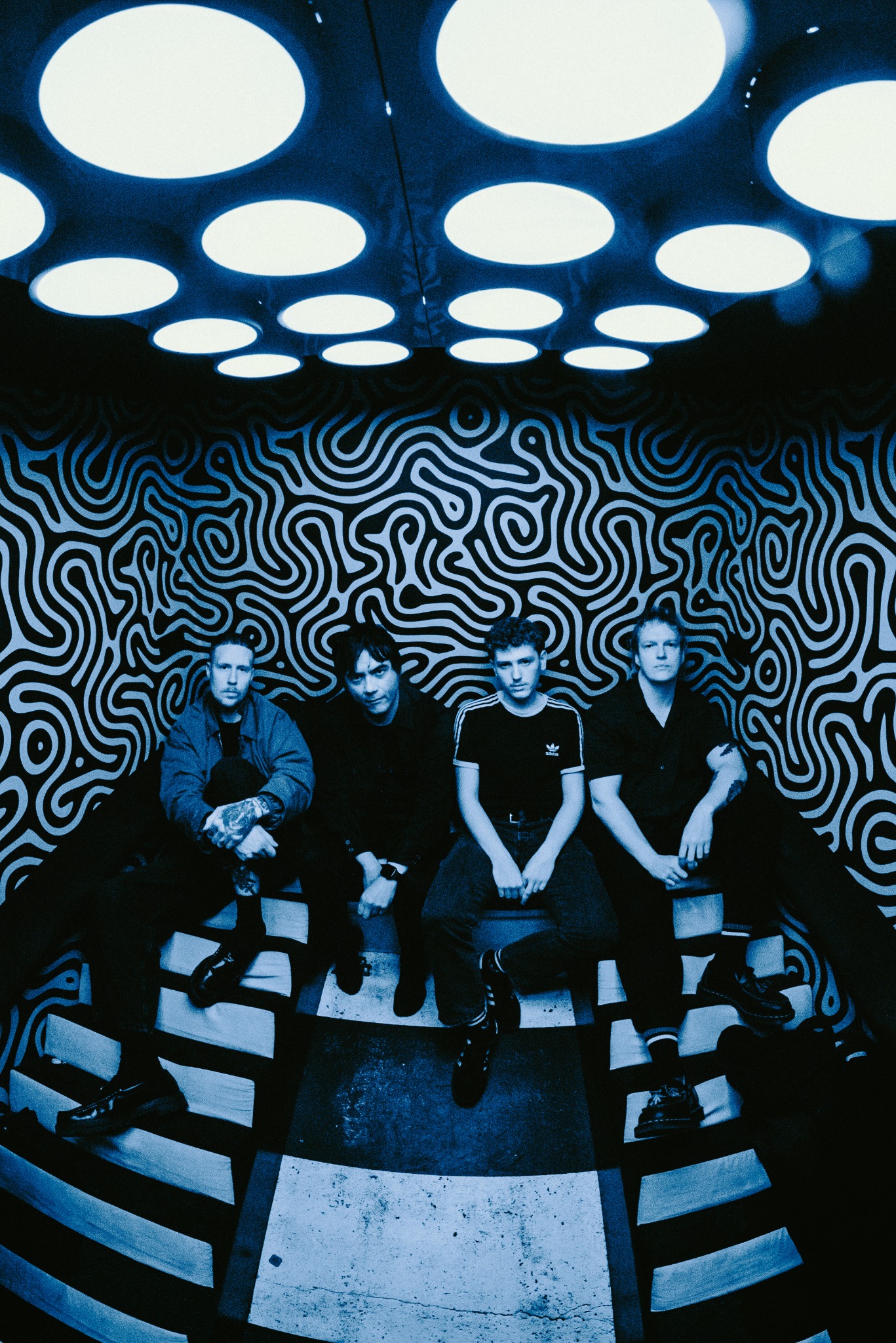 The four members of IИVDR seated beneath circular lights, photographed in blue and black.